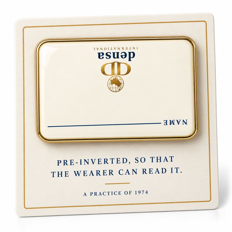 A cream enamel name badge with a gold rim, printed upside down with the Society's emblem, wordmark and a line for the wearer's name, on a card reading PRE-INVERTED, SO THAT THE WEARER CAN READ IT.